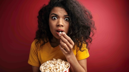 Image of shocked brunette african american woman with curly hair expressing wonder while holding popcorn bucket isolated over red background